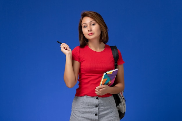 Vista frontale giovane studentessa in camicia rossa che indossa uno zaino e tenendo il quaderno su sfondo azzurro.