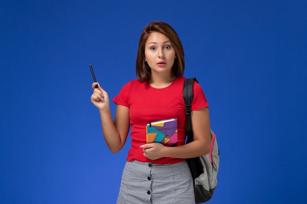 Vista frontale giovane studentessa in camicia rossa che indossa uno zaino in possesso di quaderno su sfondo blu.