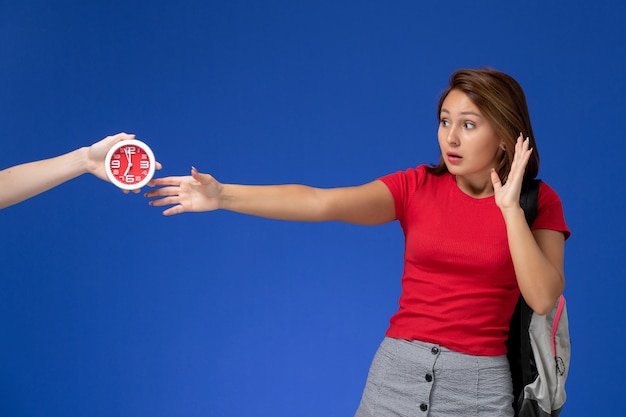 Vista frontale giovane studentessa in camicia rossa che indossa uno zaino prendendo orologi su sfondo azzurro.