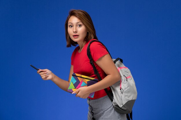 Vista frontale giovane studentessa in camicia rossa che indossa uno zaino tenendo il quaderno con la penna su sfondo azzurro.