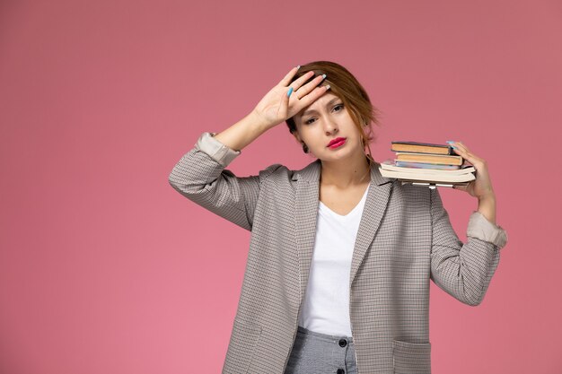 Vista frontale giovane studentessa in cappotto grigio con quaderni stanchi su sfondo rosa lezioni di studio universitario