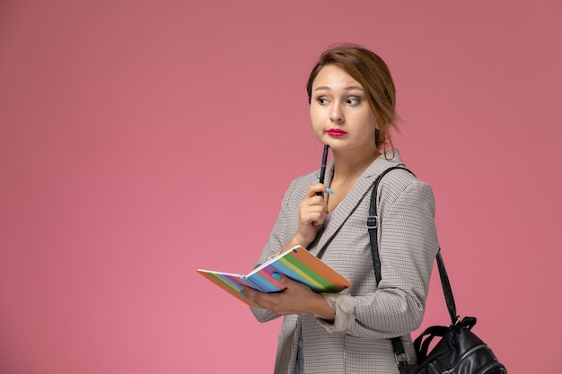 Vista frontale giovane studentessa in cappotto grigio in posa tenendo il quaderno con espressione di pensiero sullo sfondo rosa lezioni di studio universitario