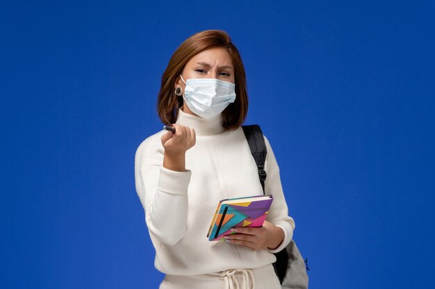 Vista frontale giovane studentessa in jersey bianco che indossa la maschera con borsa e quaderno con la penna sulla scrivania blu lezione scuola universitaria universitaria