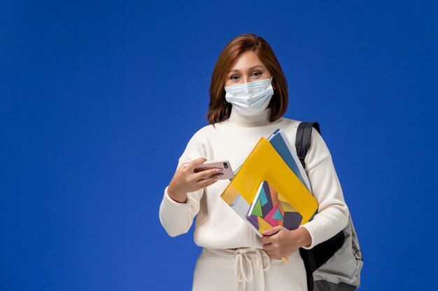 Vista frontale giovane studentessa in maglia bianca che indossa la maschera con borsa e quaderni e utilizzando il suo telefono sulla parete blu