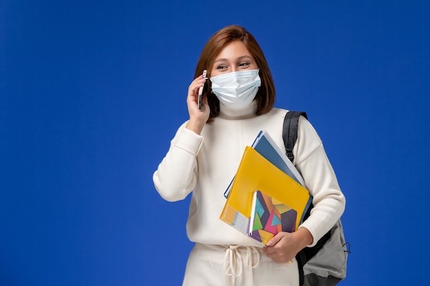 Vista frontale giovane studentessa in maglia bianca che indossa la maschera con borsa e quaderni parlando al telefono sulla parete blu