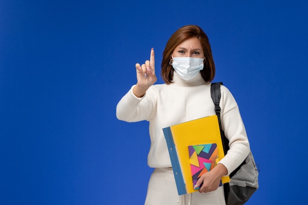 Vista frontale giovane studentessa in maglia bianca che indossa la maschera con borsa e quaderni sulla parete blu