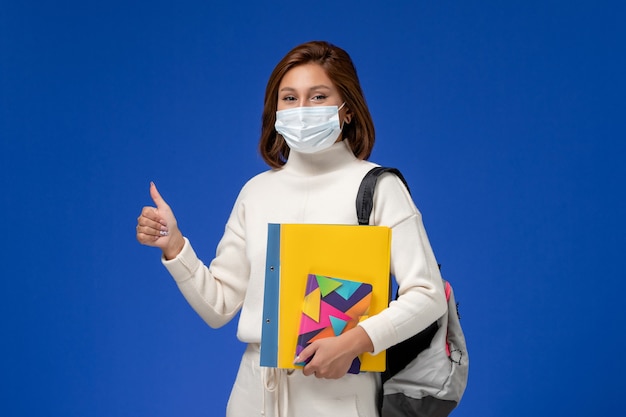 Vista frontale giovane studentessa in maglia bianca che indossa la maschera con borsa e quaderni sulla parete blu