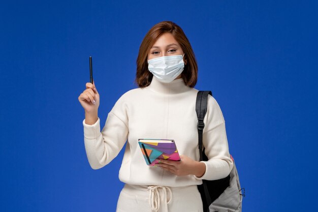 Vista frontale giovane studentessa in maglia bianca che indossa la maschera con borsa e quaderno con la penna sulla parete blu