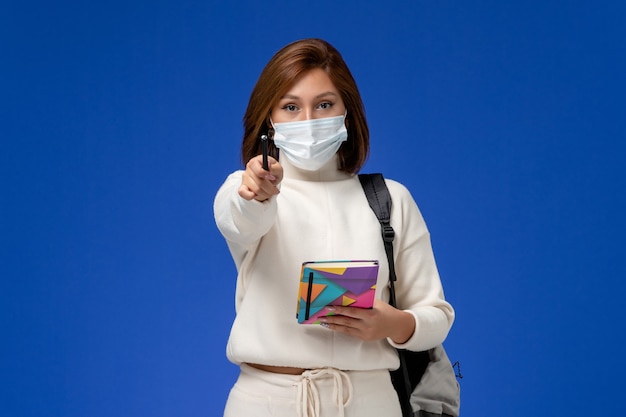 Vista frontale giovane studentessa in maglia bianca che indossa la maschera con borsa e quaderno con la penna sulla parete blu