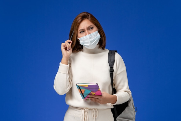 Vista frontale giovane studentessa in maglia bianca che indossa la maschera con borsa e quaderno pensando sulla parete blu