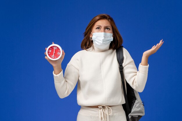 Vista frontale giovane studentessa in maglia bianca che indossa la maschera e tenendo l'orologio sulla parete blu