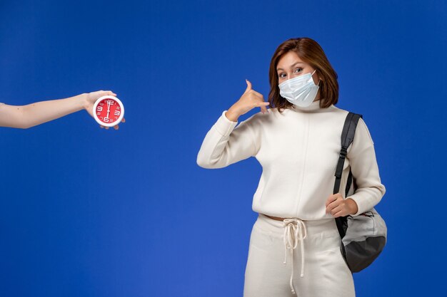 Vista frontale giovane studentessa in maglia bianca che indossa maschera e borsa sulla parete blu