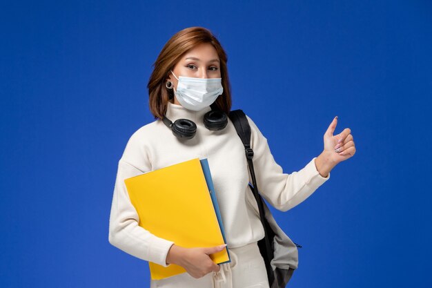 Vista frontale giovane studentessa in maglia bianca che indossa maschera e zaino tenendo i file sulla parete blu