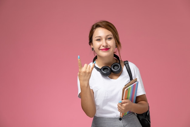 Vista frontale giovane studentessa in maglietta bianca con borsa e auricolari stile rocker in posa e sorridente su sfondo rosa lezione università college studio libro