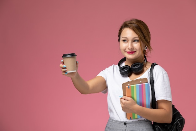 Vista frontale giovane studentessa in t-shirt bianca con borsa e auricolari in posa e sorridente che tiene caffè sul libro di studio sfondo rosa lezione università college