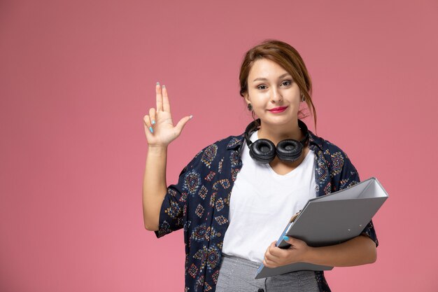 Vista frontale giovane studentessa in t-shirt bianca e pantaloni grigi con documento grigio nelle sue mani sullo sfondo rosa studente lezioni universitarie