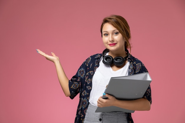 Vista frontale giovane studentessa in t-shirt bianca e pantaloni grigi con documento grigio nelle sue mani sullo sfondo rosa studente lezioni universitarie