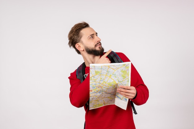 Vista frontale giovane turista maschio con lo zaino esplorando la mappa sul muro bianco aereo città vacanza emozione colore umano itinerario turistico