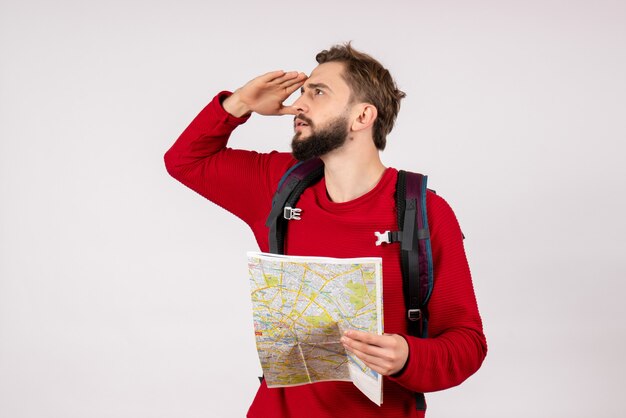 Vista frontale giovane turista maschio con zaino esplorando la mappa sul percorso del turismo umano di emozione di vacanza di città aereo muro bianco