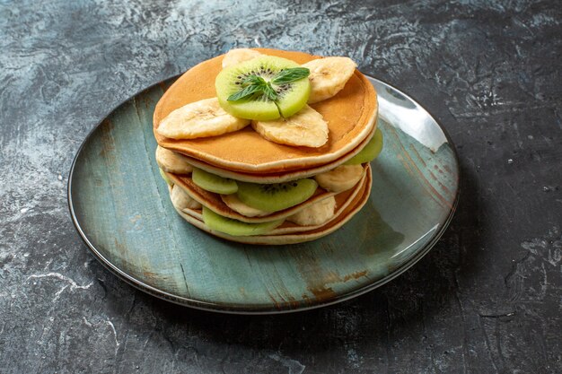 Vista frontale gustose frittelle con kiwi a fette e banane su superficie scura frutta dolce colazione torta zucchero colore dolce