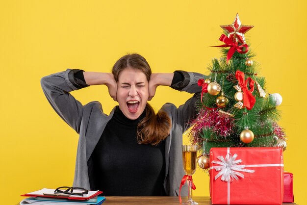 Vista frontale ha gridato la ragazza che si siede allo scrittorio che copre le orecchie con le sue mani albero di Natale e cocktail di regali