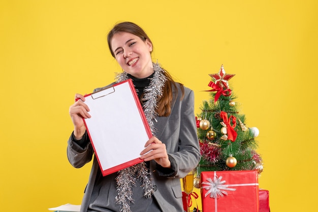 Vista frontale ha sorriso la ragazza con i documenti in piedi vicino all'albero di Natale e al cocktail dei regali