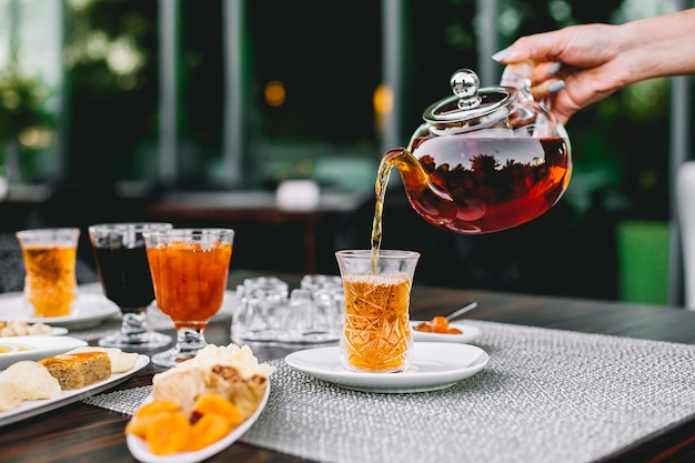 Vista frontale la ragazza versa il tè dalla teiera in un bicchiere di armoud con marmellata e dolci sul tavolo