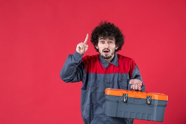 Vista frontale lavoratore maschio con cassetta degli attrezzi su sfondo rosso foto colore lavoratore meccanico casa strumento lavoro emozione uniforme