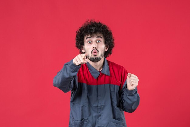 Vista frontale lavoratore maschio in uniforme che punta su sfondo rosso colore uniforme strumento lavoratore meccanico strumento lavoro casa foto