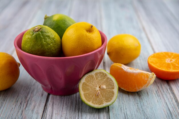 Vista frontale limette con limoni in una ciotola e fette d'arancia su sfondo grigio