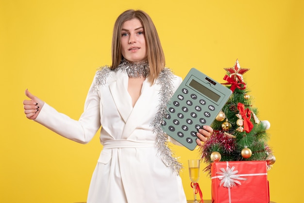 Vista frontale medico femminile in piedi e tenendo il calcolatore su sfondo giallo con albero di Natale e confezioni regalo