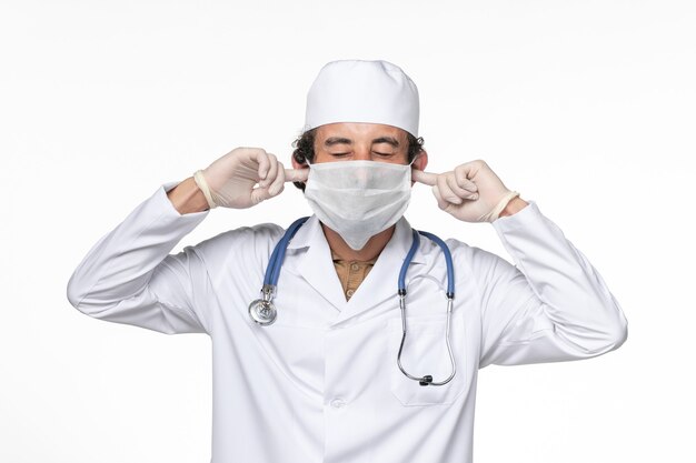 Vista frontale medico maschio in tuta medica con maschera come protezione da covid- chiudendo le orecchie sul muro bianco virus del coronavirus medicina pandemica