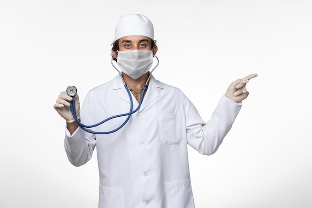 Vista frontale medico maschio in tuta medica e indossa una maschera come protezione da covid- utilizzando uno stetoscopio sulla scrivania bianca malattia covid- pandemia