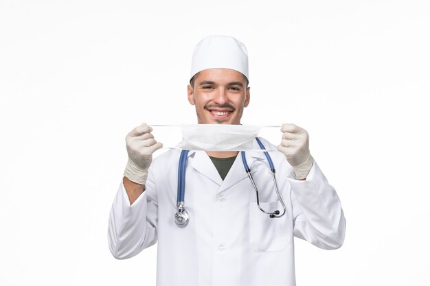 Vista frontale medico maschio in tuta medica e indossa una maschera contro il coronavirus sorridente sul muro bianco virus covid - malattia pandemica