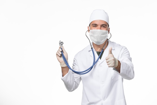 Vista frontale medico maschio in tuta medica e maschera da indossare a causa di covid- utilizzando uno stetoscopio sorridente sul muro bianco covid- malattia da virus pandemico
