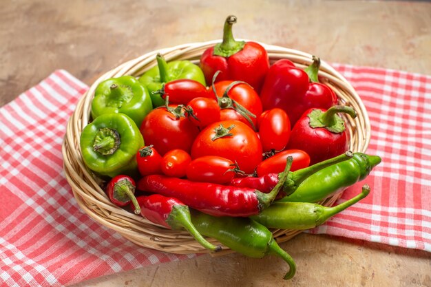 Vista frontale peperoni verdi e rossi peperoncini pomodori in cesto di vimini e asciugamano da cucina su sfondo ambrato