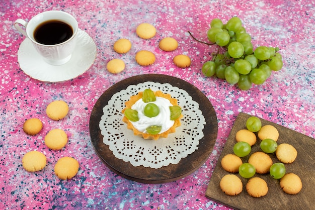 Vista frontale piccola torta con crema tazza di biscotti da tè e insieme con uva verde sulla frutta torta superficie luminosa