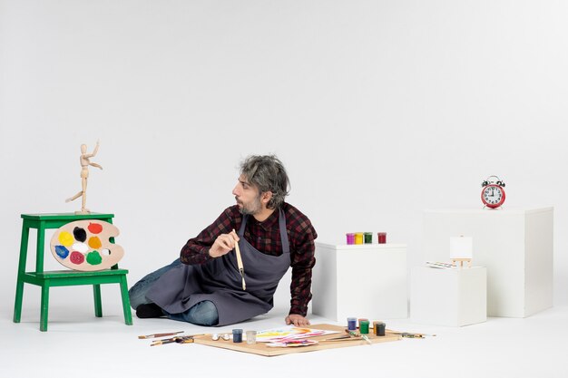 Vista frontale pittore maschio seduto con vernici e nappe per disegnare sullo sfondo bianco arte disegno immagine artista pittura a colori