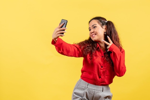 Vista frontale ragazza in camicetta rossa con capelli carini che si fanno selfie su sfondo giallo bambina ragazza gioventù innocenza colore bambino