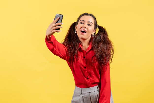 Vista frontale ragazza in camicetta rossa con capelli carini che si fanno selfie su sfondo giallo bambina ragazza gioventù innocenza colore bambino