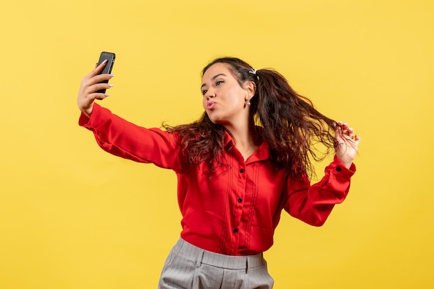 Vista frontale ragazza in camicetta rossa con capelli carini che si fanno selfie sulla scrivania gialla ragazzino ragazza gioventù innocenza colore bambino