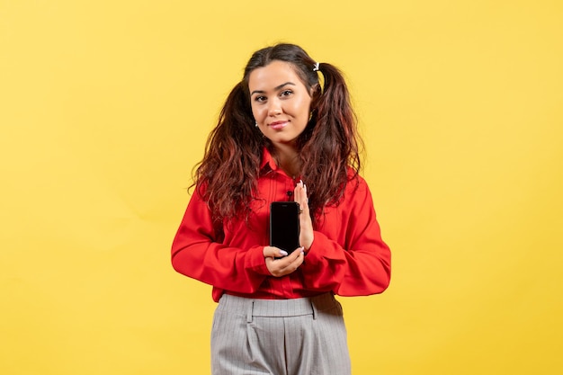 Vista frontale ragazza in camicetta rossa con capelli carini che tiene il telefono sulla scrivania gialla ragazzino ragazza gioventù innocenza colore bambino