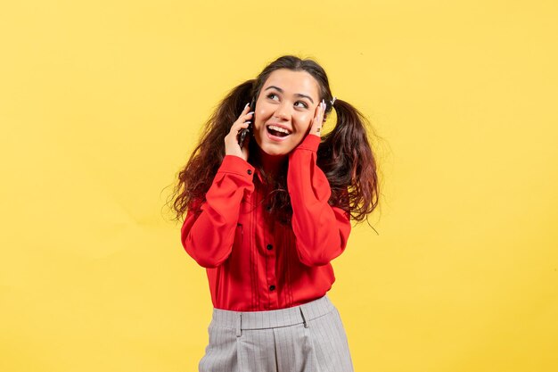 Vista frontale ragazza in camicetta rossa con capelli carini parlando al telefono su sfondo giallo colore bambino ragazza bambino gioventù innocenza