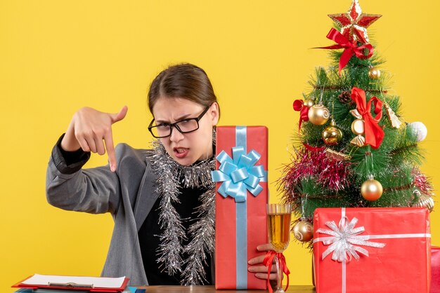 Vista frontale ragazza non gratificata con gli occhiali seduti al tavolo indicato con il dito verso il basso albero di Natale e regali cocktail