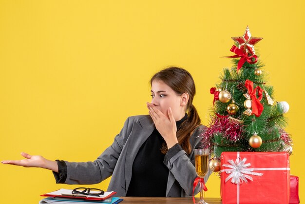 Vista frontale ragazza sorpresa seduta al tavolo che mostra qualcosa vicino all'albero di Natale e regali cocktail