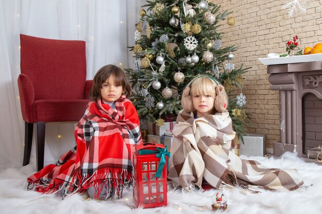 Vista frontale ragazzini carini seduti intorno all'albero di natale e regali nella loro casa colore foto capodanno infanzia bambino natale