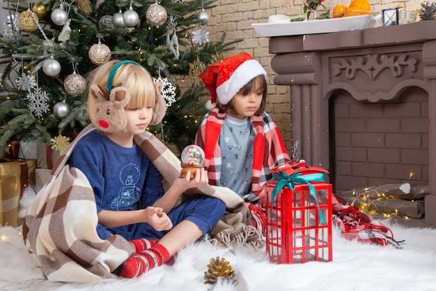 Vista frontale ragazzini carini seduti intorno all'albero di Natale e regali nella loro casa