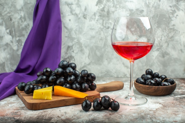 Vista frontale ravvicinata del delizioso grappolo di uva nera fresca e formaggio su tagliere di legno e in una pentola marrone un bicchiere di vino su sfondo di colore misto