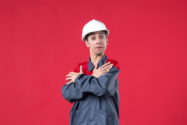 Vista frontale ravvicinata del giovane costruttore in uniforme che indossa un elmetto e si sente sorpreso sul muro rosso isolato