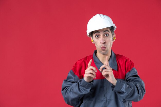 Vista frontale ravvicinata del giovane costruttore scioccato in uniforme con elmetto e con i tappi per le orecchie che puntano verso l'alto sul muro rosso isolato